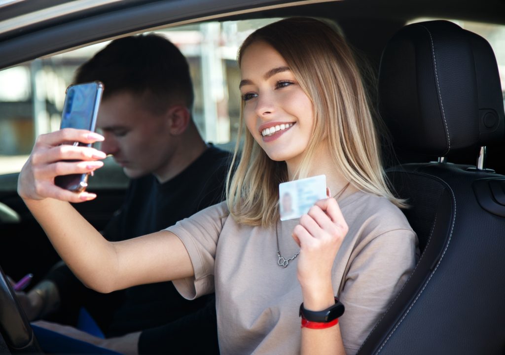 Happy smiling girl takes a selfie with a new drivers license, sitting next to an instructor